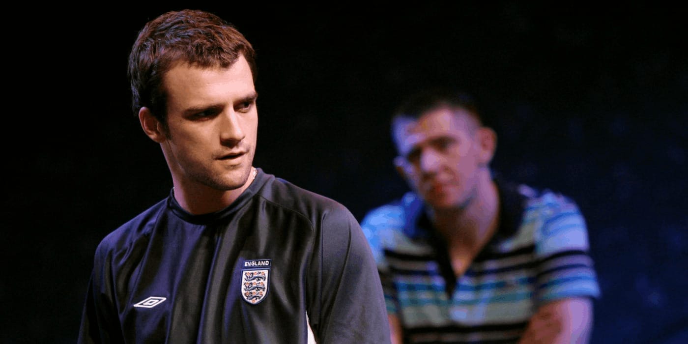 A man in a grey England football top that has the three lions emblem on with a man in a stripy polo shirt sat behind him