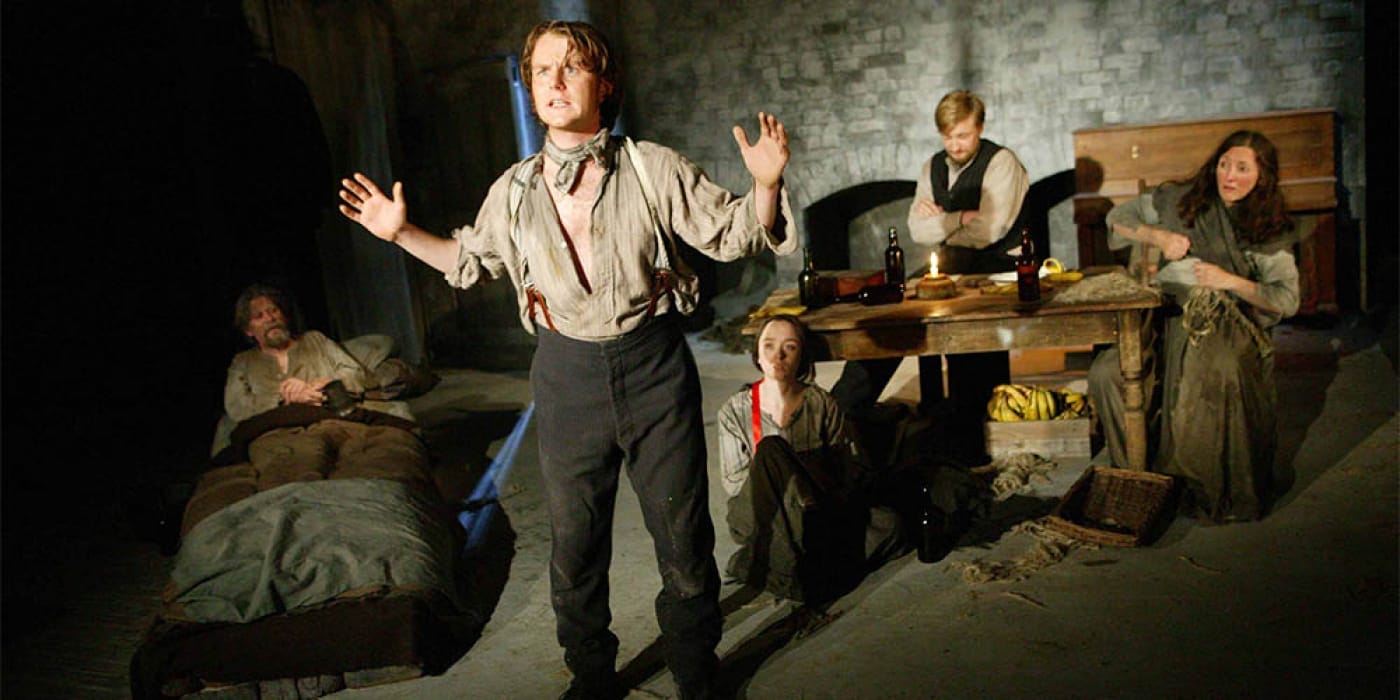 A man stood in a beige half unbuttoned shirt with his arms out and black trousers . Behind him is a family of two women and two men.