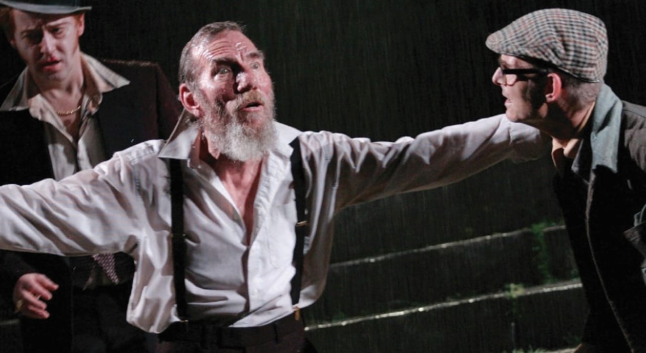 A man in a white shirt and black suspenders talking with his arms wide to the side to a man in a flat cap.