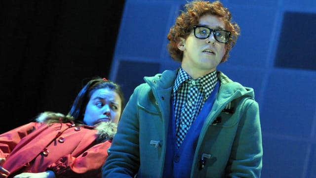 A man with ginger curly hair is stood wearing glasses, a gingham shirt, blue button up cardigan and green duffle coat. behind them is a girl with brunette hair leaning back on a chair wearing a red parker coat.