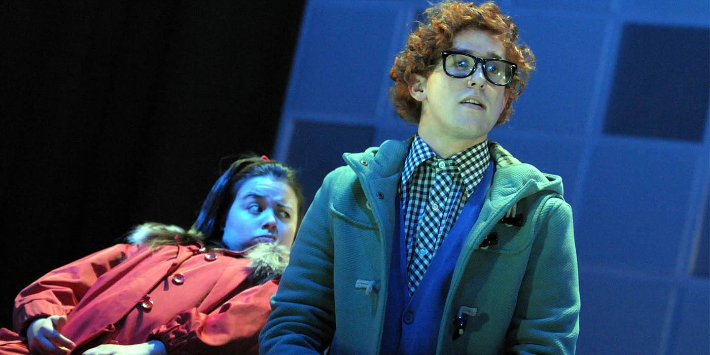 A man with ginger curly hair is stood wearing glasses, a gingham shirt, blue button up cardigan and green duffle coat. behind them is a girl with brunette hair leaning back on a chair wearing a red parker coat.