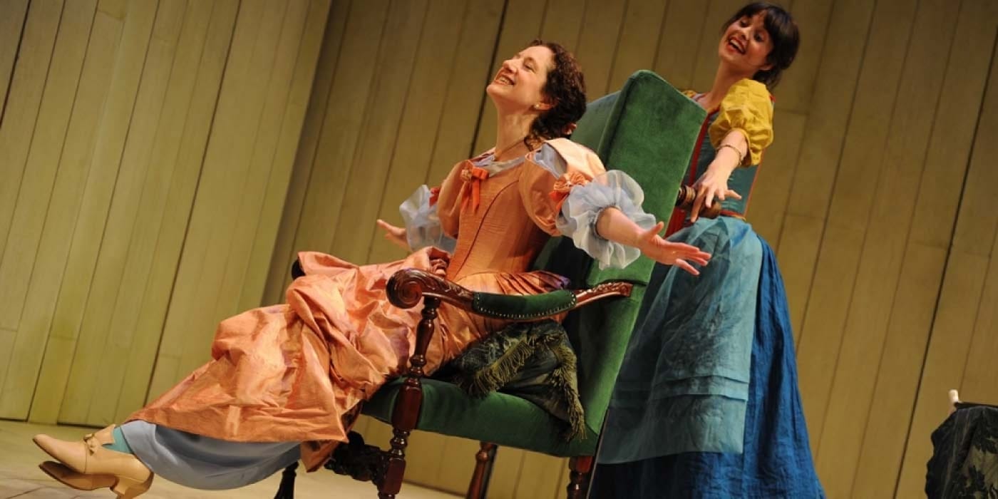 A woman in a pink dress sat on a chair with her arms spread. There is a woman behind her holding the back of the chair wearing a blue dress