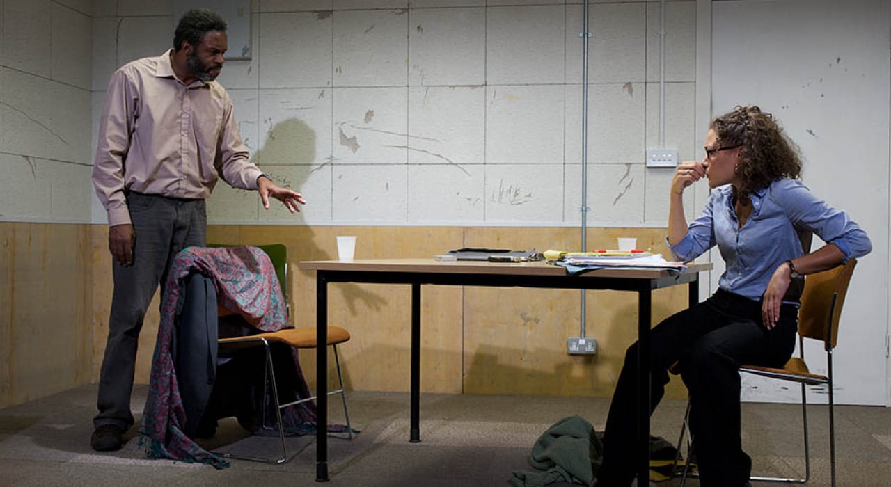 A man stood behind a chair talking to a woman who is sat across from him at the table