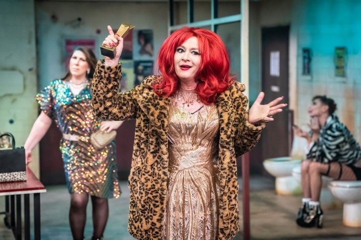 A person with long red hair holding a gold trophy. They are wearing a leopard print fur coat and a nude gold sequin dress. in the back ground there us someone sat on the toilet and another person stood in a sequin dress.