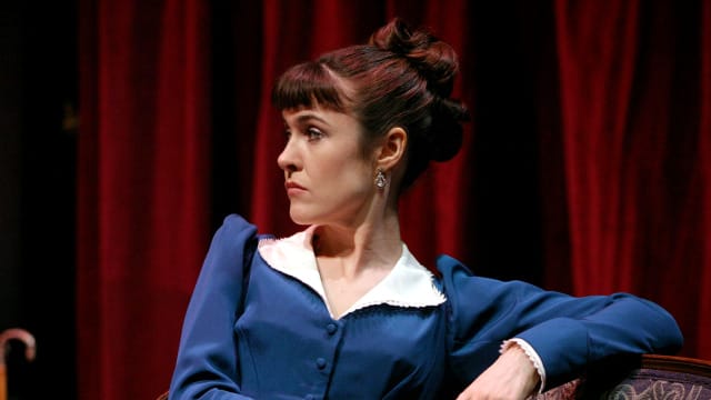 A woman in a bless dress and white collar sat on a chair. Her elbow is propped on the back support. She has brown hair tied into a bun and has a full fringe. Her facial expressions shows shes not pleased.