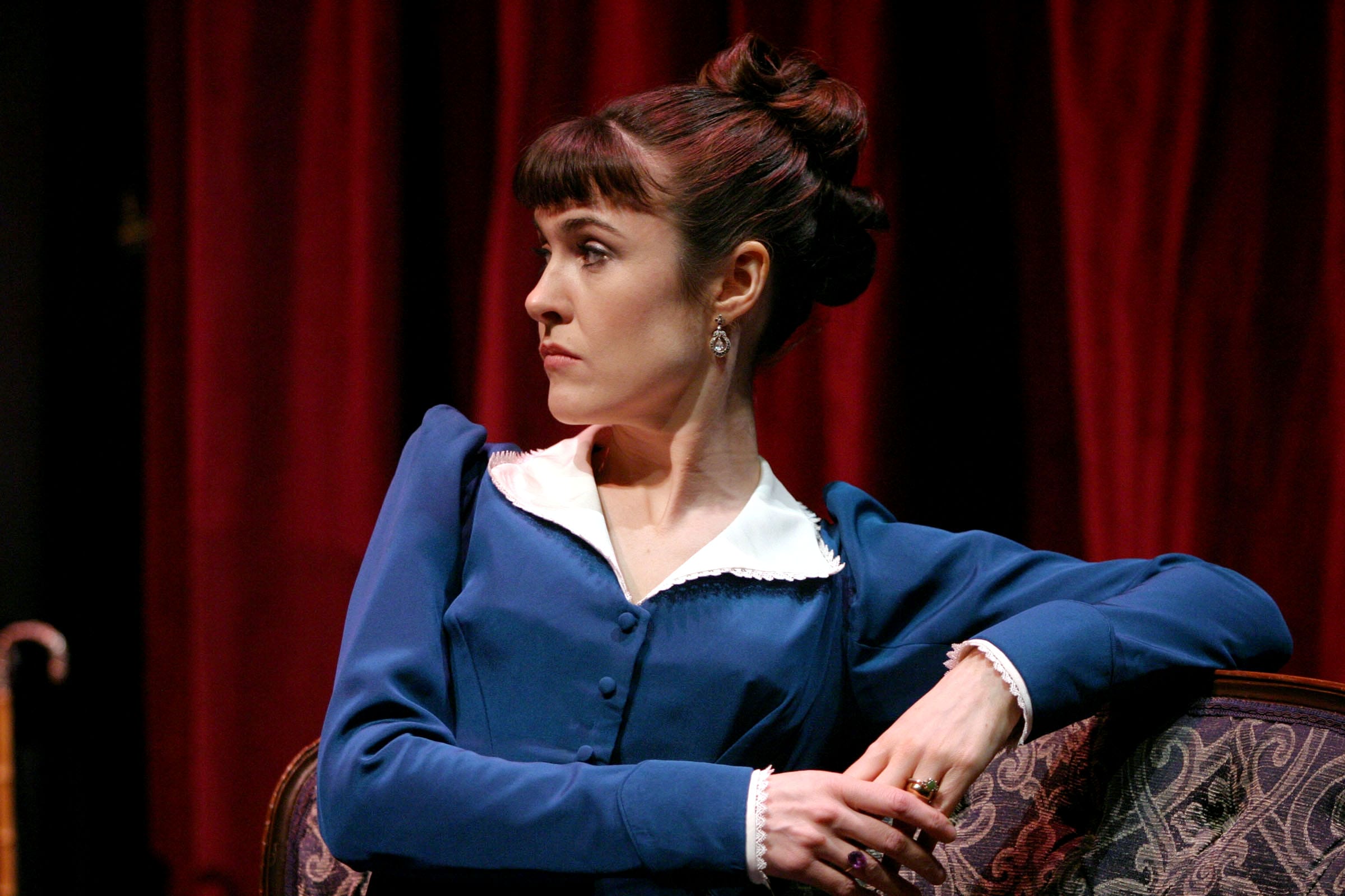 A woman in a bless dress and white collar sat on a chair. Her elbow is propped on the back support. She has brown hair tied into a bun and has a full fringe. Her facial expressions shows shes not pleased.