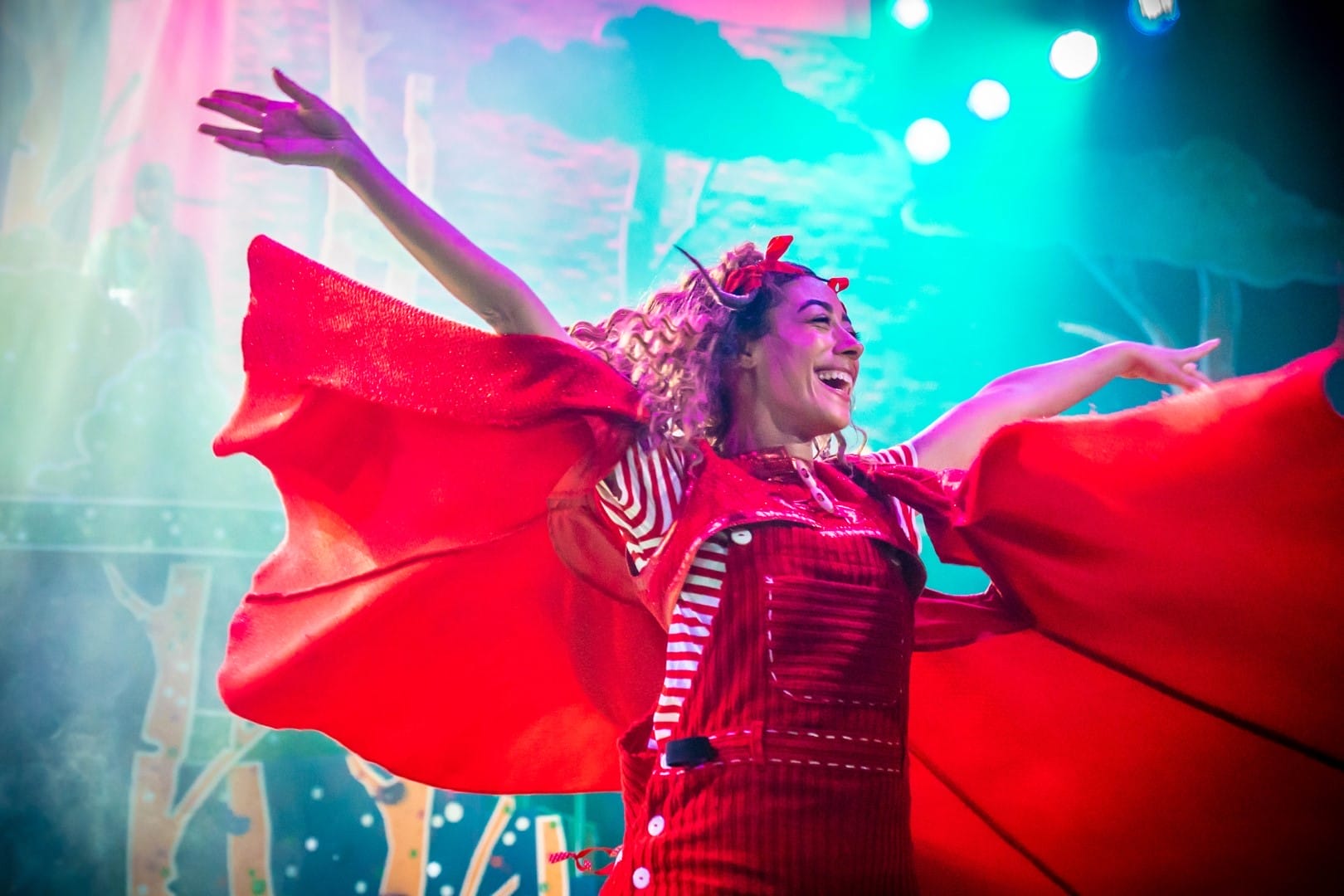 A Photograph from the panto 2022 showing Red Riding Hood, wearing a large red cape with her arms spread out wide and a smile on her face.