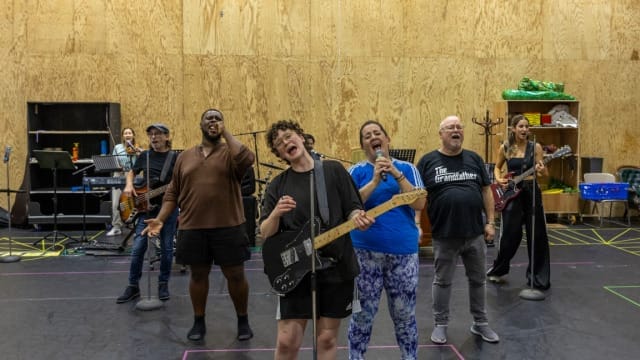 7 actors in rehearsals for Rapunzel, they are standing in a triangle formation singing into microphones, some are also playing instruments (keyboard and guitars)