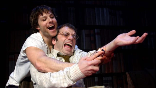 A man attempting to burn his arm with a lighter and a man holding his arm up to assist him doing so