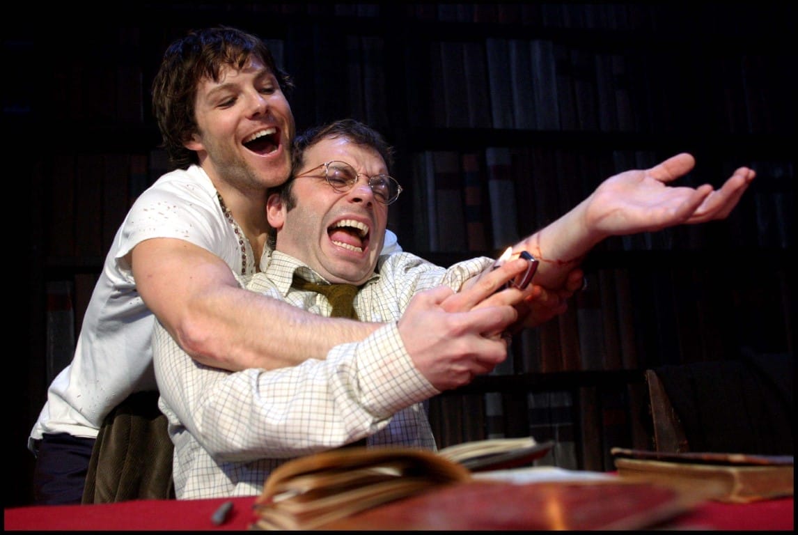 A man attempting to burn his arm with a lighter and a man holding his arm up to assist him doing so