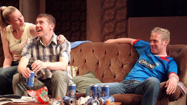 A brown sofa with a woman sat on the arm. She is wearing a yellow vest and has her arm around the shoulders of a man who is sat on the sofa. He has a checkered blue short sleeve shirt on and is drinking a can of lager. On the opposite side of the sofa is another man in a blue football shirt with carling across the center of it. Infront of them is a table with multiple empty cans on and sweet wrappers.