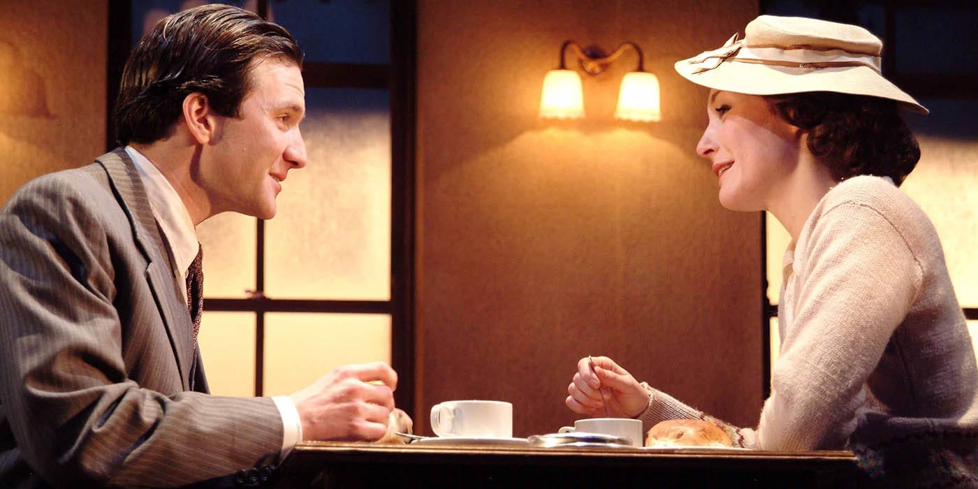 A woman and man sat across from eachother having coffee. The woman is wearing a white dress and a white hat. they are staring into eachothers eyes with a soft smile.