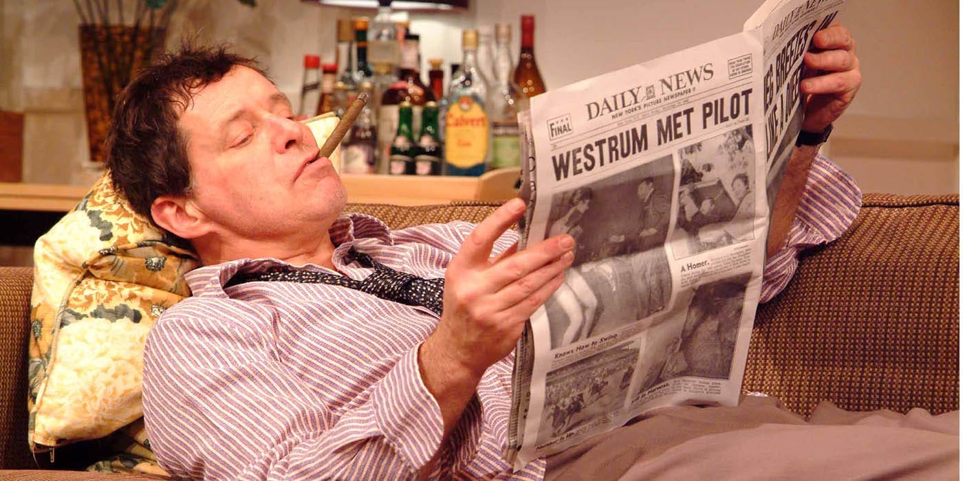 A man lying on the sofa with a newspaper in his hands and a cigar in his mouth. In the background is multiple bottles of alcohol