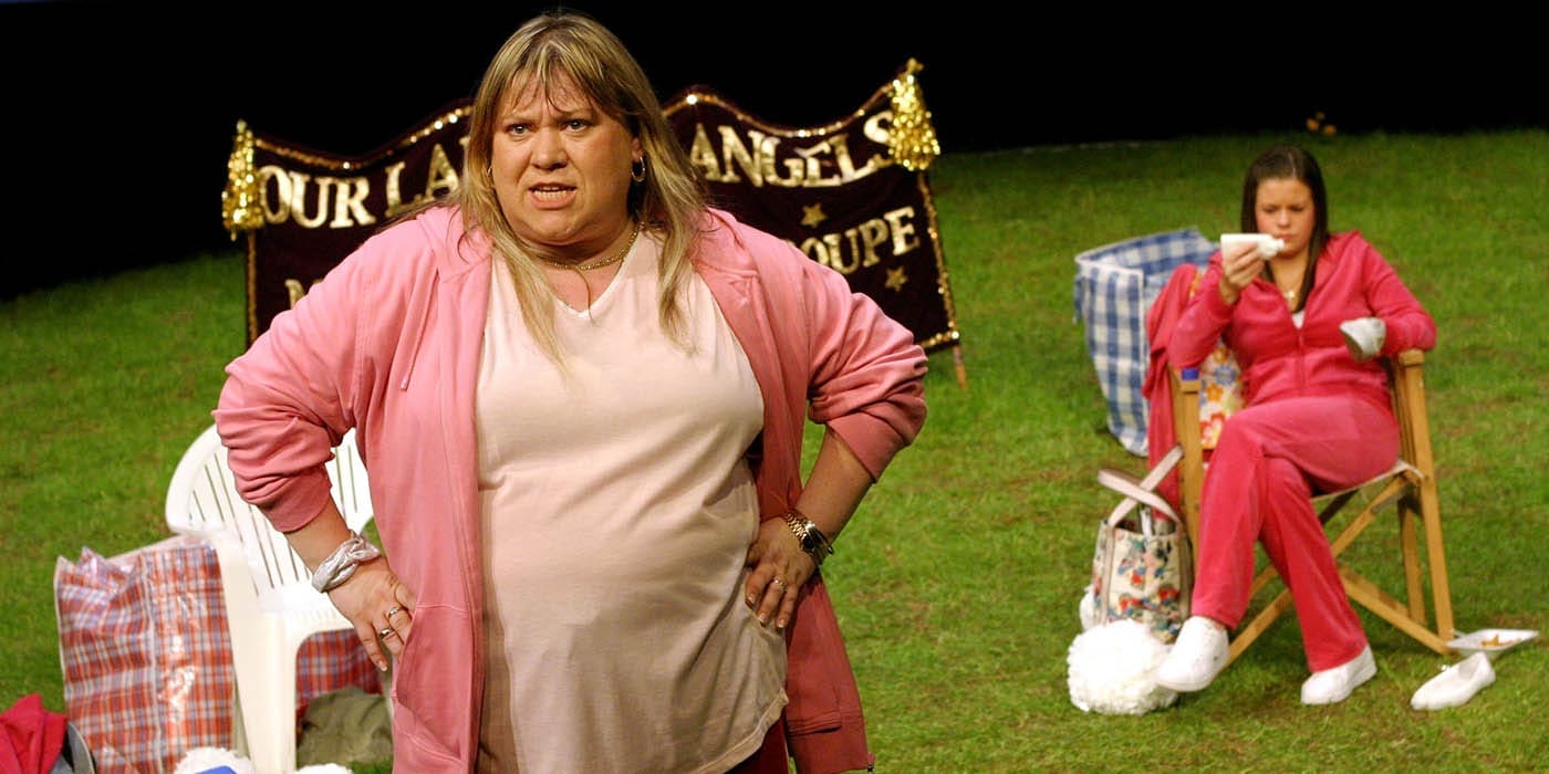 A woman in a light pink top and pink cardigan stood with her hands on her hips. In the background is a grass field and another woman in a pink tracksuit sat on a fold out chair cleaning her shoes.